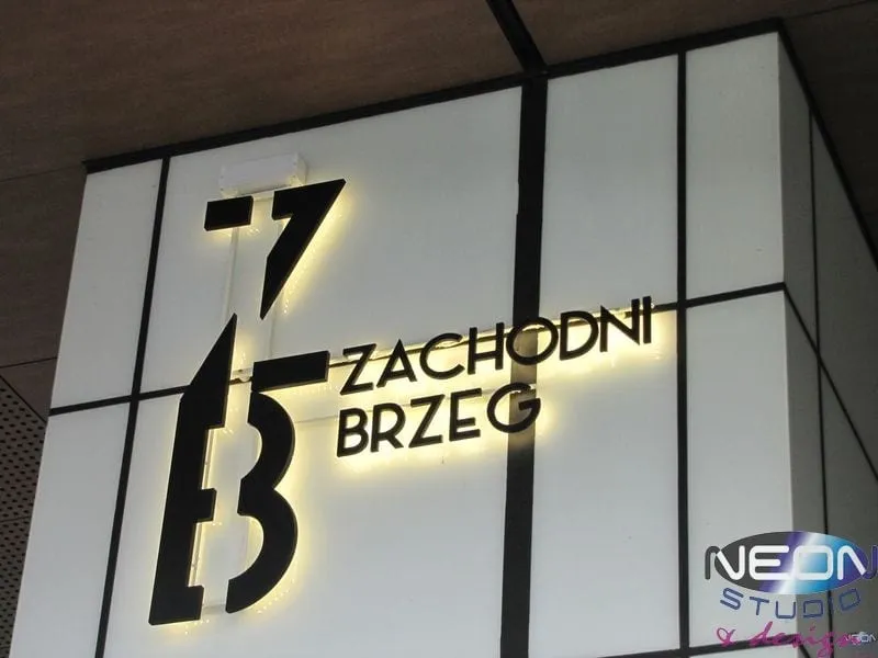 Litery z efektem halo — ciemne lico i poświata na jasnej okładzinie elewacji, znak „Zachodni Brzeg”